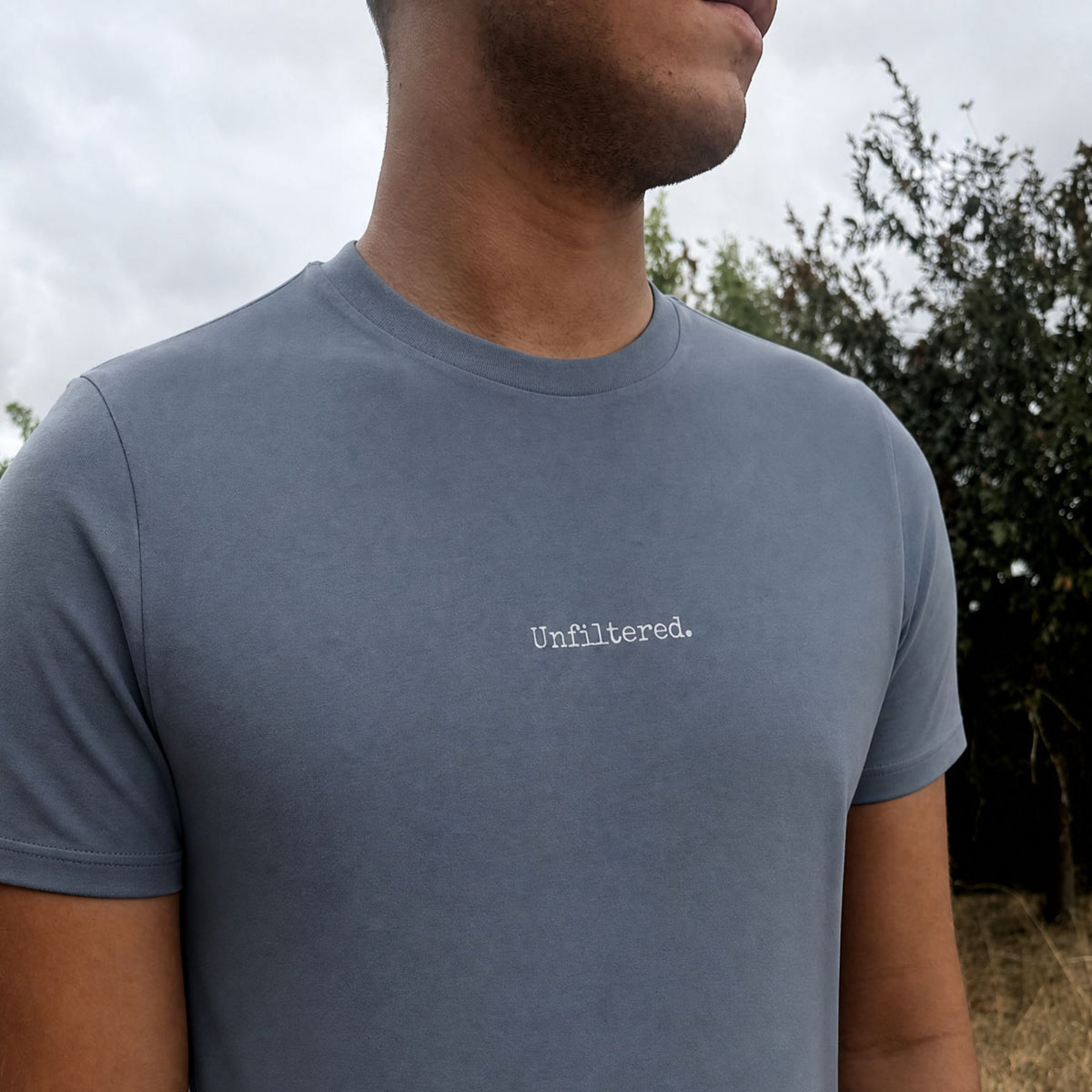A man wearing a slate blue crewneck t-shirt with the word 'Unfiltered.' printed in white typewriter font on the chest, photographed outdoors against a cloudy sky and trees.