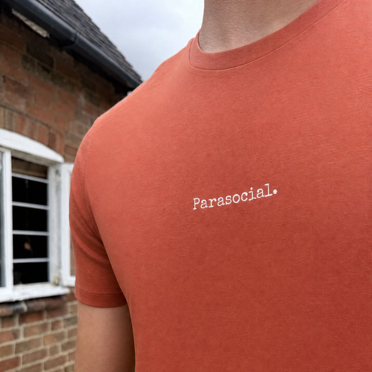 A close-up of a person wearing a terracotta orange crewneck t-shirt with 'Parasocial.' printed in white typewriter font on the chest, set against a blurred brick building background.