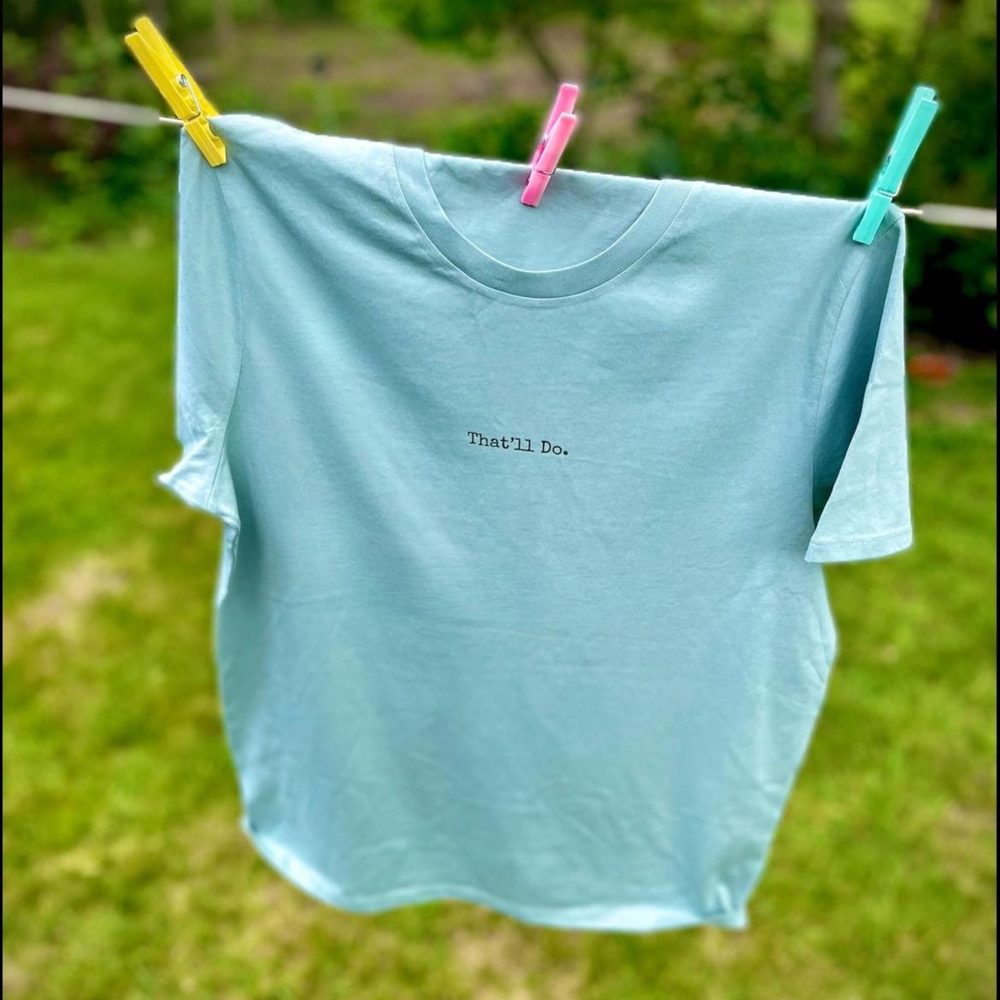 Light blue t-shirt with 'That'll Do.' text hanging on a clothesline with green grass in the background.