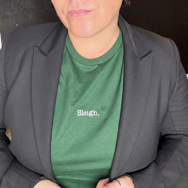 A person wearing a  black blazer over a green crewneck t-shirt with the word 'Sleigh.' embroidered in white on the chest.