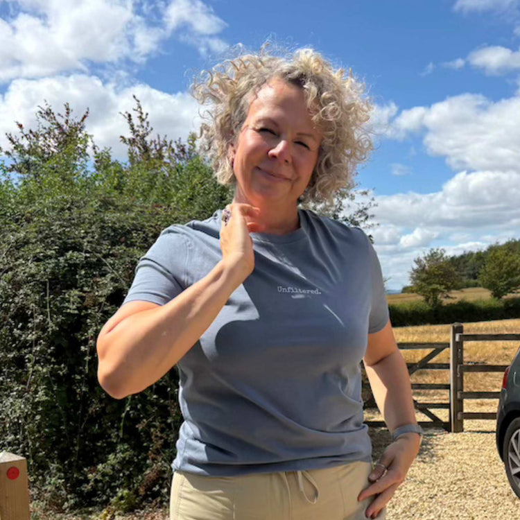 A woman with curly blonde hair wearing a slate blue t-shirt that says unfiltered and beige trousers 