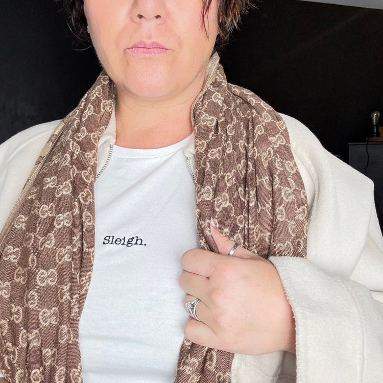 A person wearing a brown and beige monogram-patterned scarf over a white t-shirt with the word 'Sleigh.' printed on the chest, paired with a cream-colored zip-up jacket.