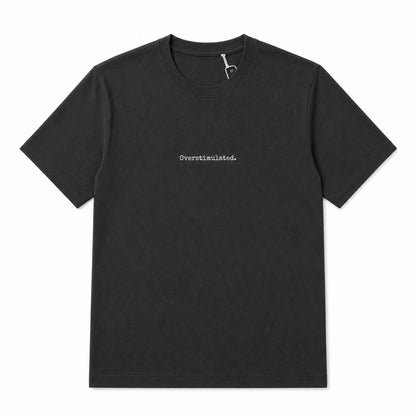 A black short-sleeve crewneck t-shirt with the word 'Overstimulated.' printed in small white serif text on the center chest, displayed flat against a solid white background.