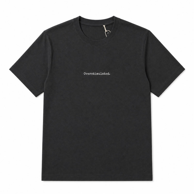 A black short-sleeve crewneck t-shirt with the word 'Overstimulated.' printed in small white serif text on the center chest, displayed flat against a solid white background.