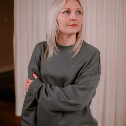 Person wearing a dark green sweatshirt with a logo, standing against a neutral background