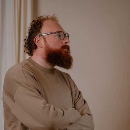 Man with a beard and glasses wearing a beige sweater against a plain background