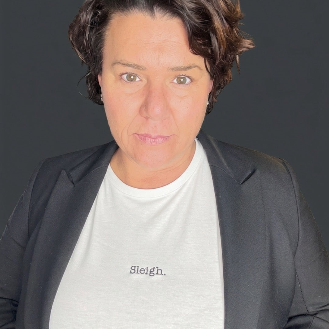 A close-up portrait of a woman wearing a light gray crewneck t-shirt with the word 'Sleigh.' printed in black script on the chest, layered under a dark gray blazer, against a dark gray background.