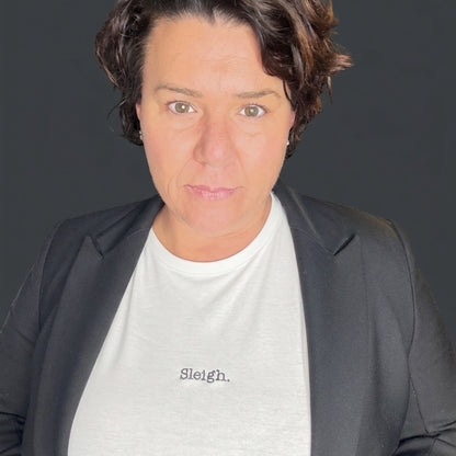 A close-up portrait of a woman wearing a light gray crewneck t-shirt with the word 'Sleigh.' printed in black script on the chest, layered under a dark gray blazer, against a dark gray background.