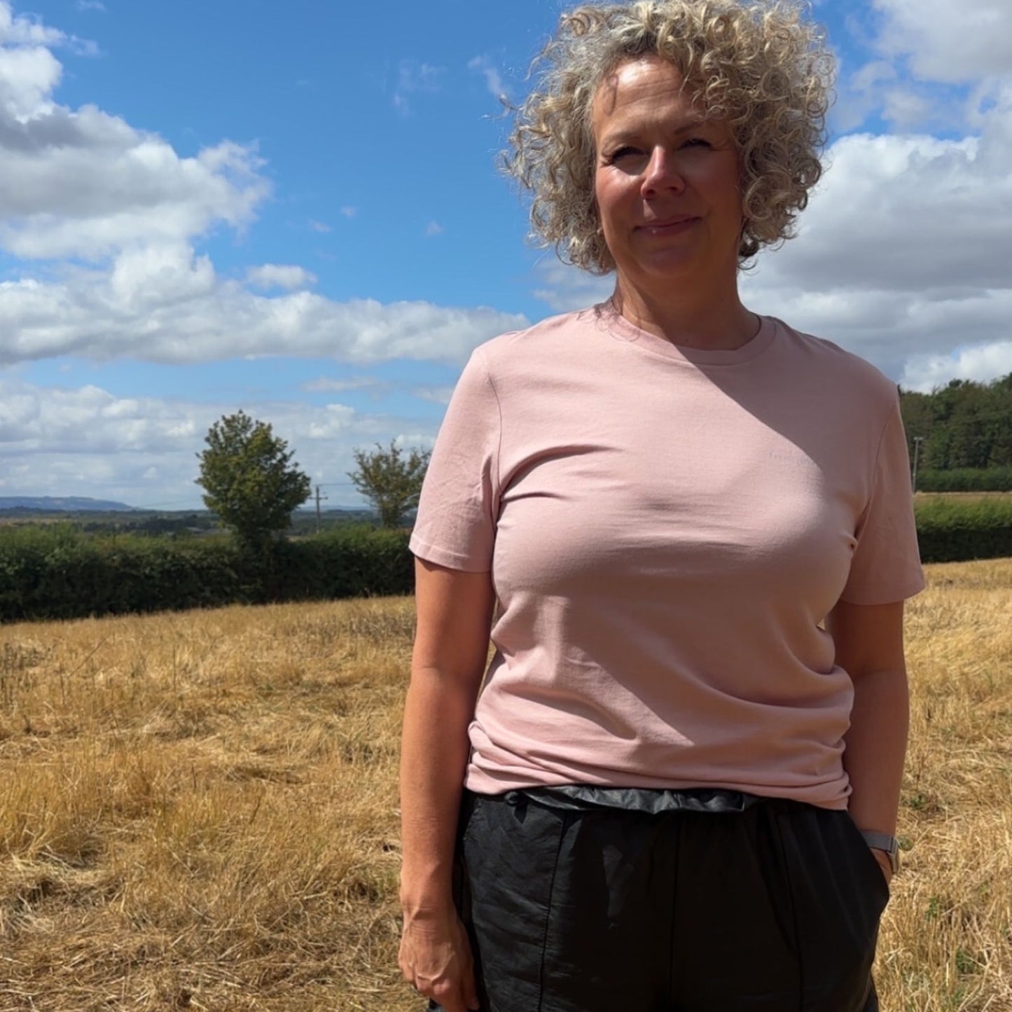 A woman with curly blonde hair stands outdoors in a field under a blue sky with clouds, wearing a light pink short-sleeve crewneck t-shirt and dark grey pants.