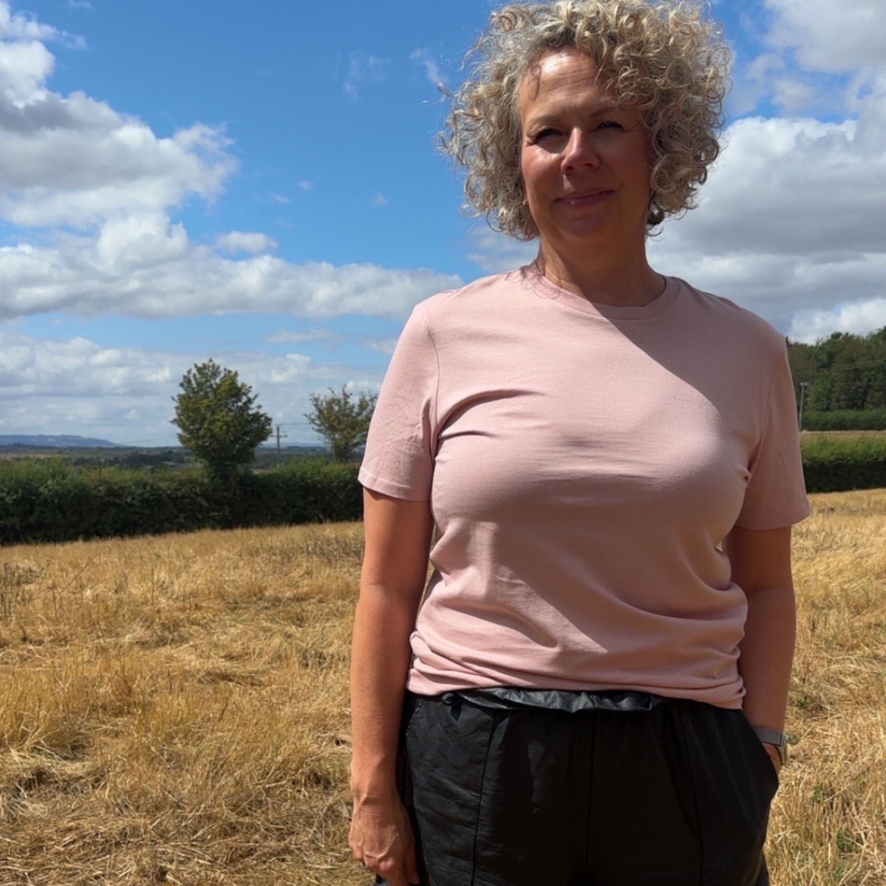 A woman with curly blonde hair stands outdoors in a field under a blue sky with clouds, wearing a light pink short-sleeve crewneck t-shirt and dark grey pants.