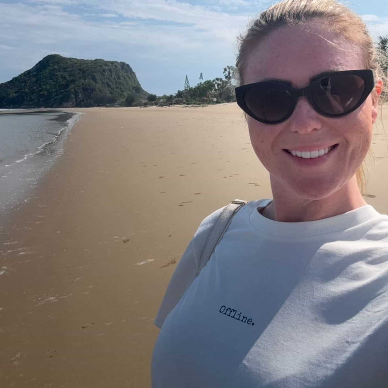 Person wearing sunglasses and a 'Offline' shirt on a beach with mountains in the background