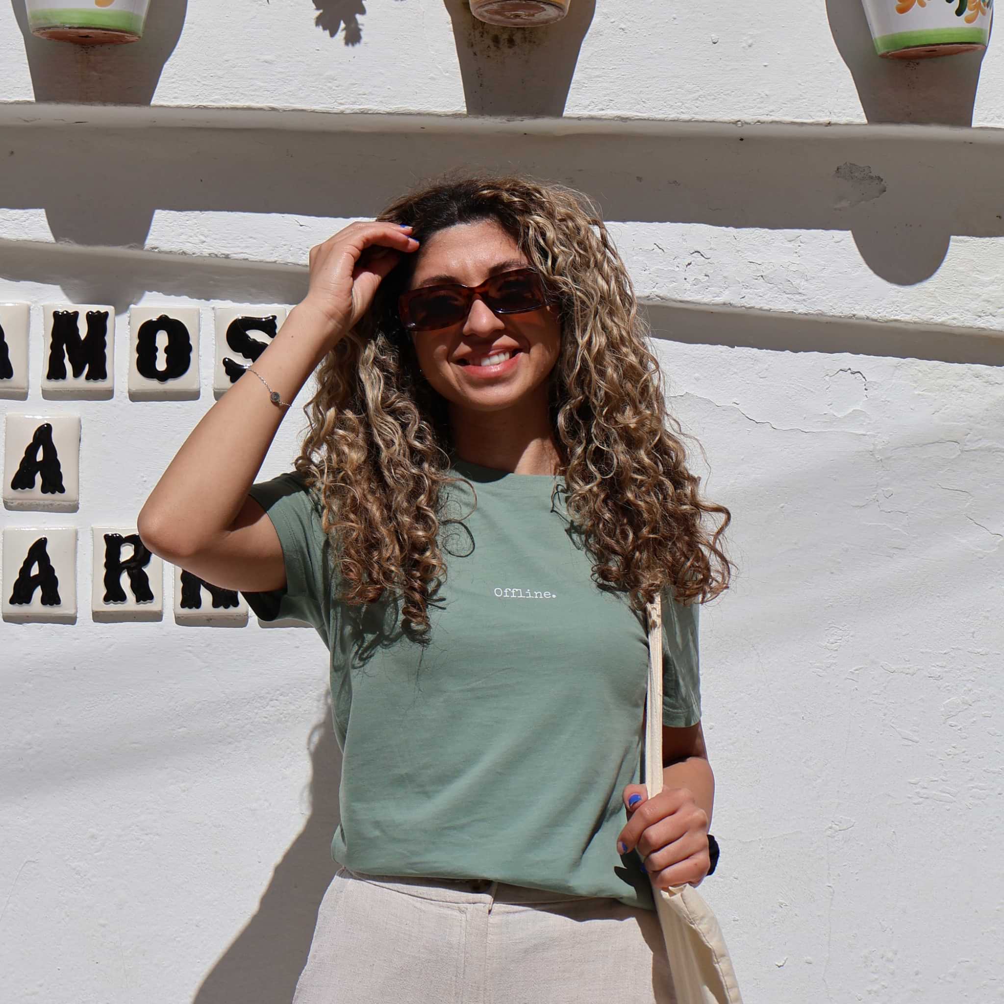 Woman with curly hair wearing sunglasses and a green t-shirt against a white wall with decorative tiles.