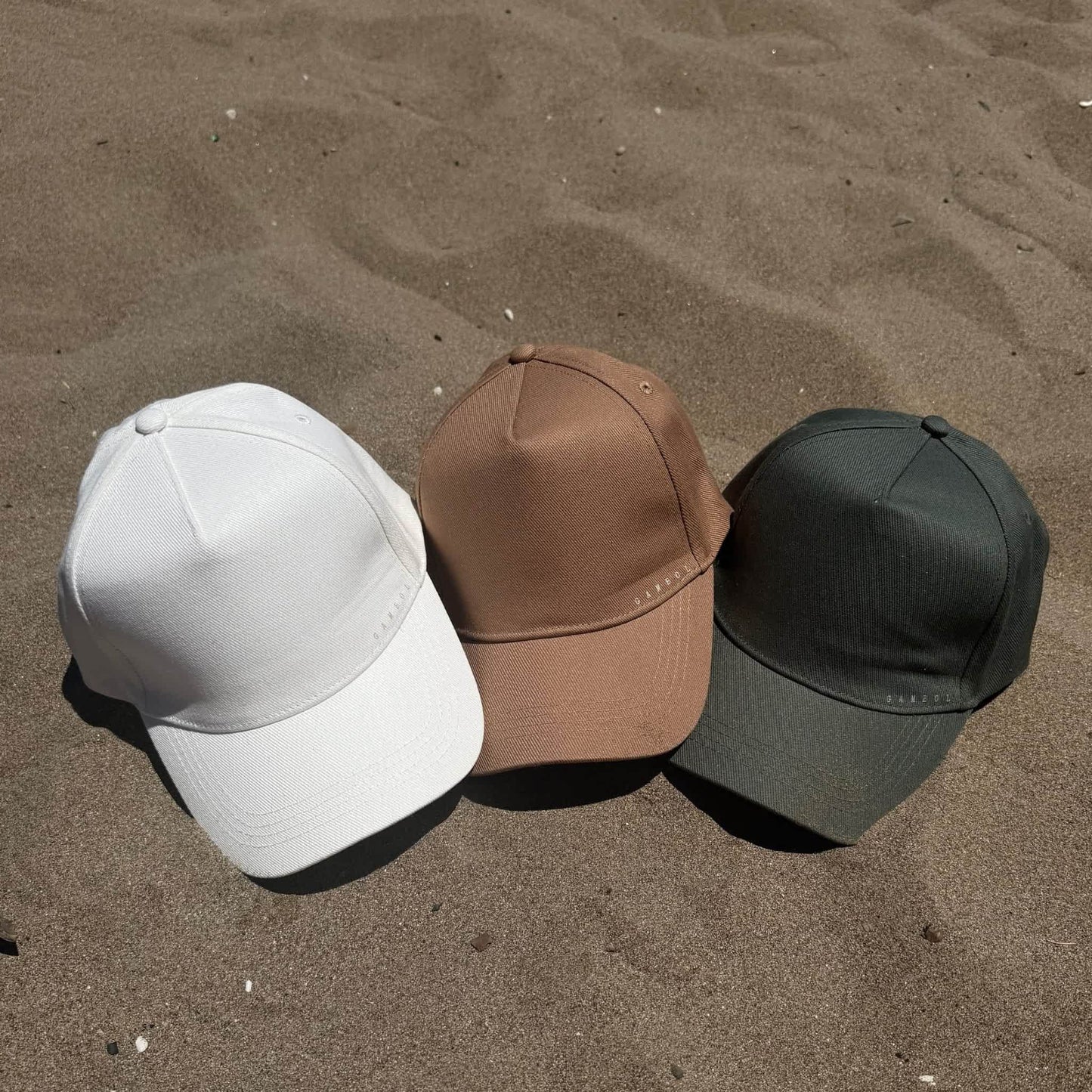 Three baseball caps in white, brown, and green on a sandy surface