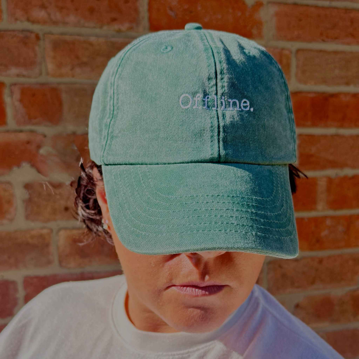 A person wearing a muted teal baseball cap with the word 'Offline.' embroidered in white on the front, photographed against a red brick wall.