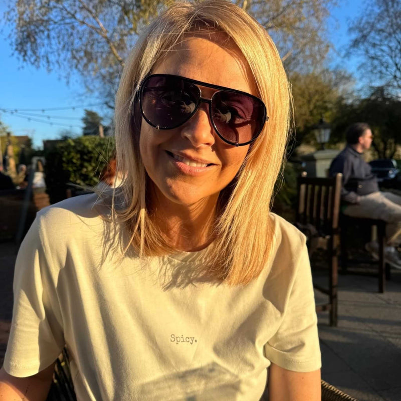 Woman wearing sunglasses and a light-colored t-shirt with 'Spicy' text, sitting outdoors.