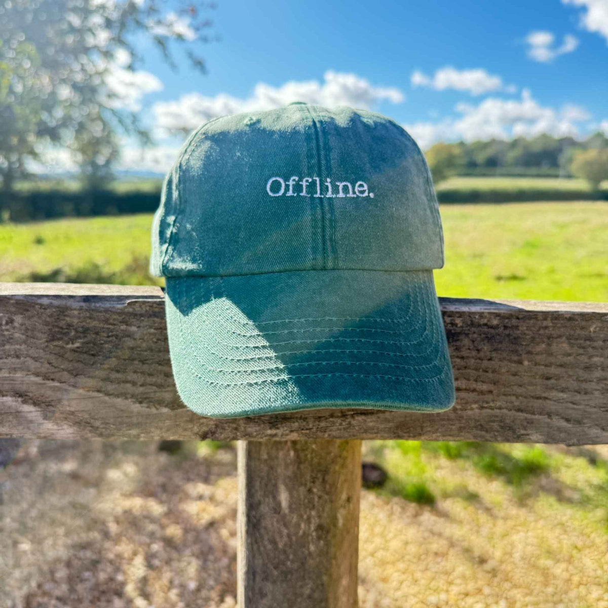 A green baseball cap with the word 'Offline.' embroidered on the front, resting on a wooden fence outdoors with a blurred field and blue sky in the background.