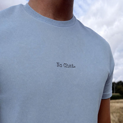 A close-up of a person wearing a light blue crewneck t-shirt with the text 'No Chat.' printed in black typewriter font on the chest, set against an outdoor background of cloudy sky and grass.