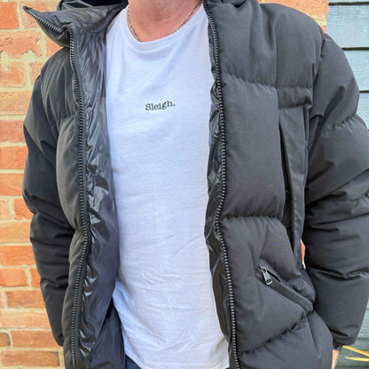 A person wearing a black puffer jacket over a white t-shirt with the word 'Sleigh.' printed on the chest, standing in front of a brick wall.
