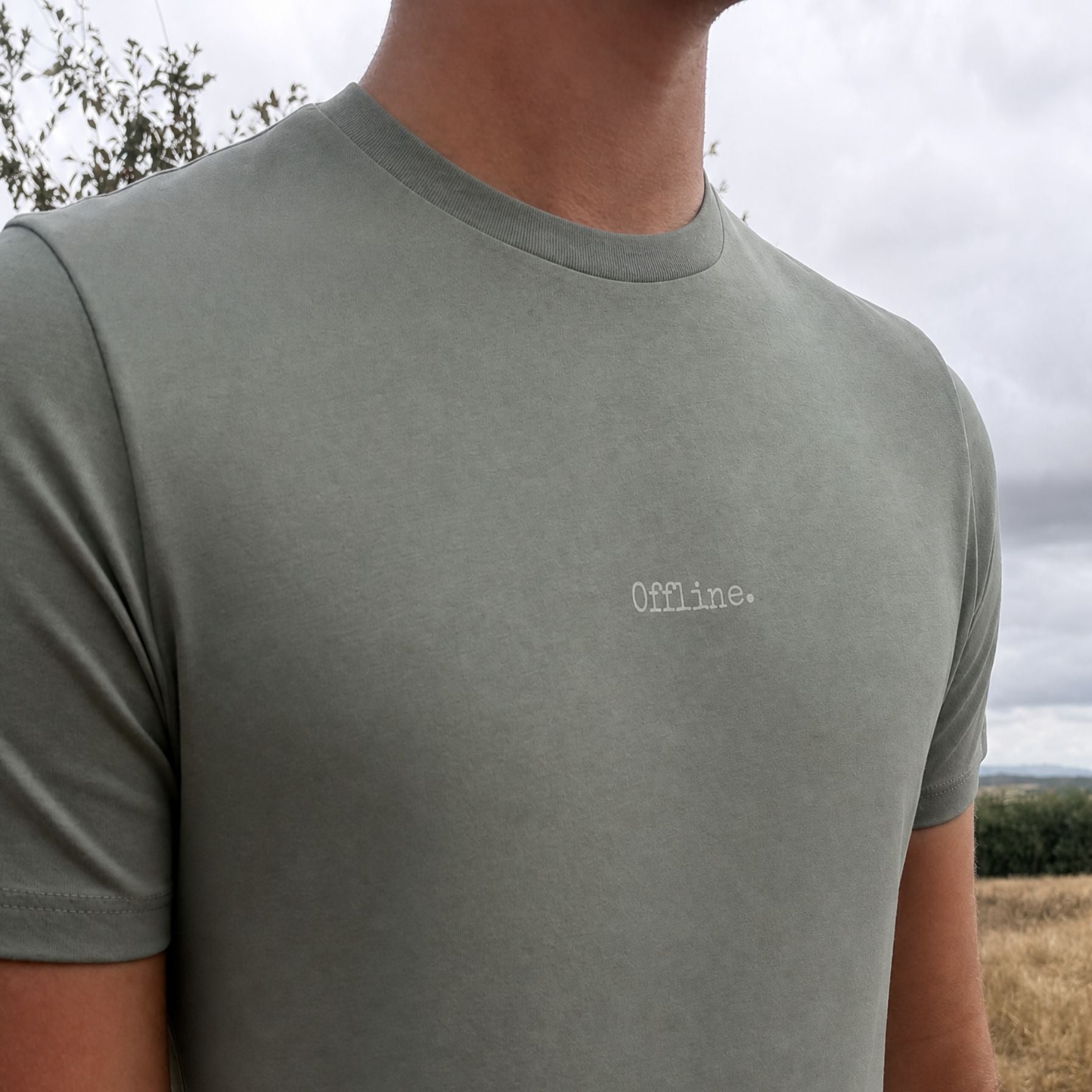 A close-up of a person wearing a muted sage green t-shirt with the word 'Offline.' embroidered in white on the chest, set against an outdoor background of cloudy sky and dry grass.