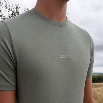A close-up of a person wearing a muted sage green t-shirt with the word 'Offline.' embroidered in white on the chest, set against an outdoor background of cloudy sky and dry grass.