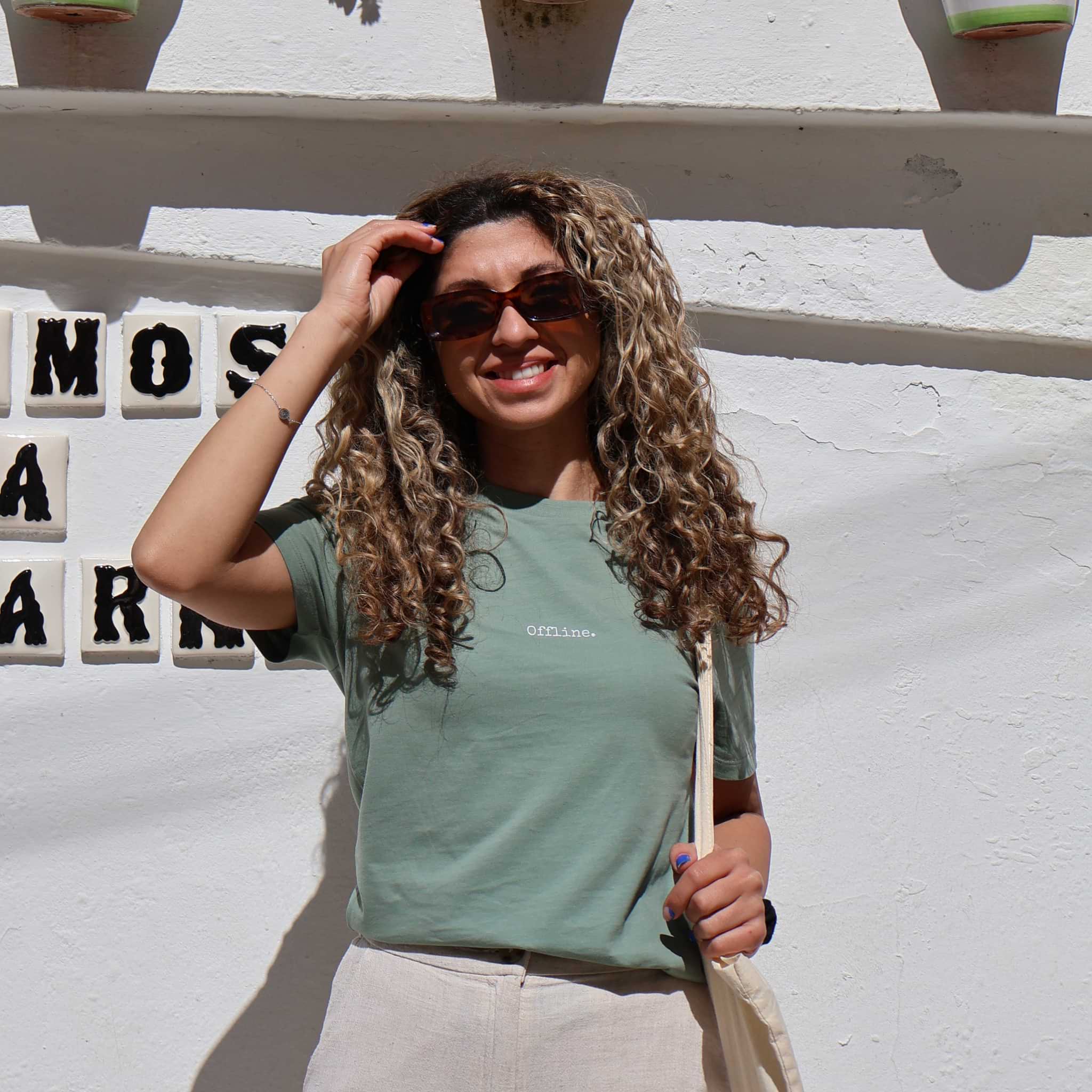 Woman with curly hair wearing sunglasses and a green t-shirt against a white wall with decorative letters.
