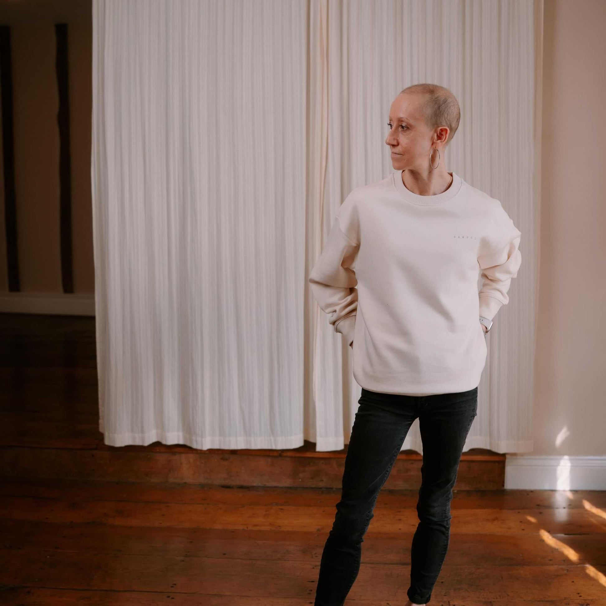 Person wearing a white sweater and black pants standing in a room with wooden floor and white curtains.