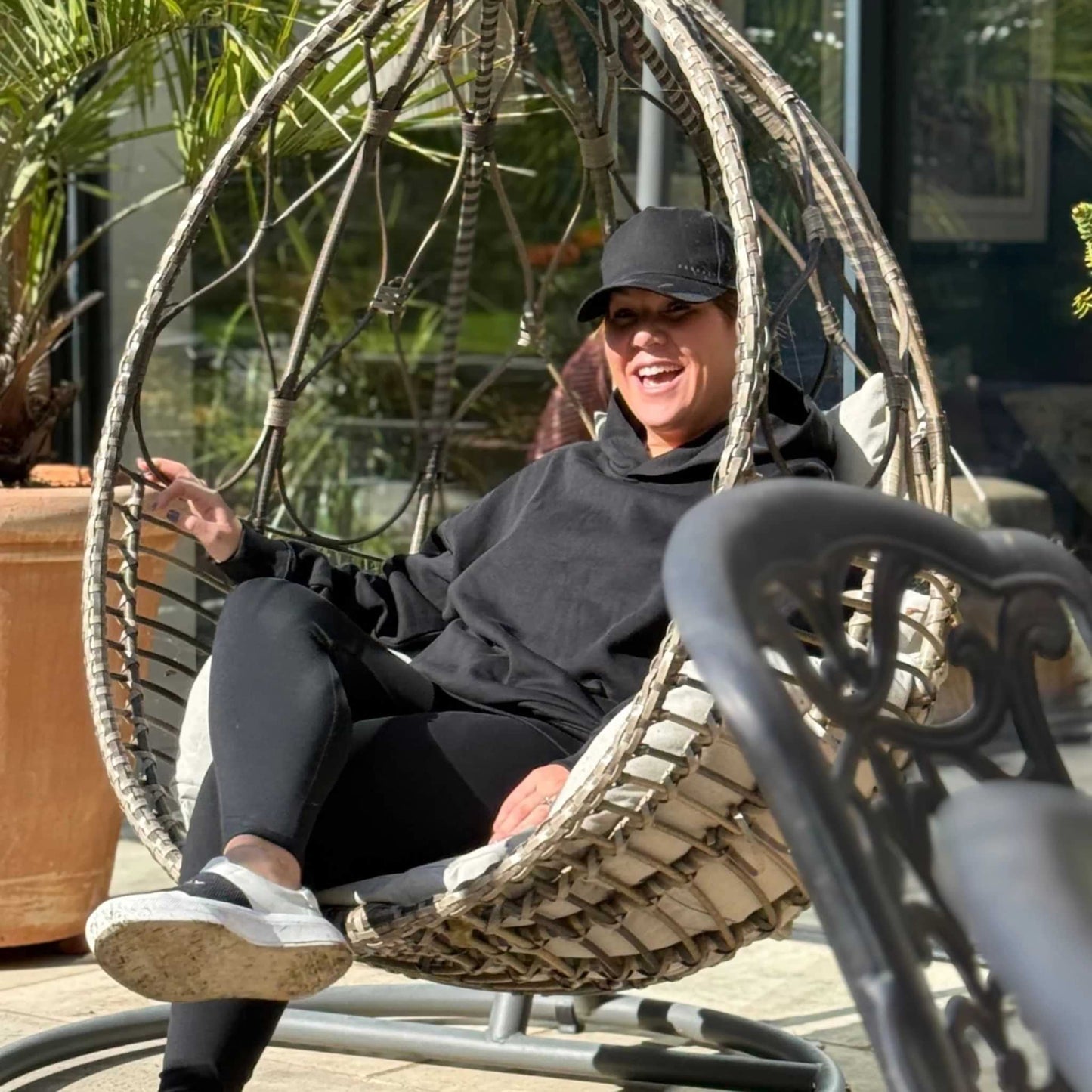 Person sitting in a hanging chair outdoors with plants in the background wearing black cap