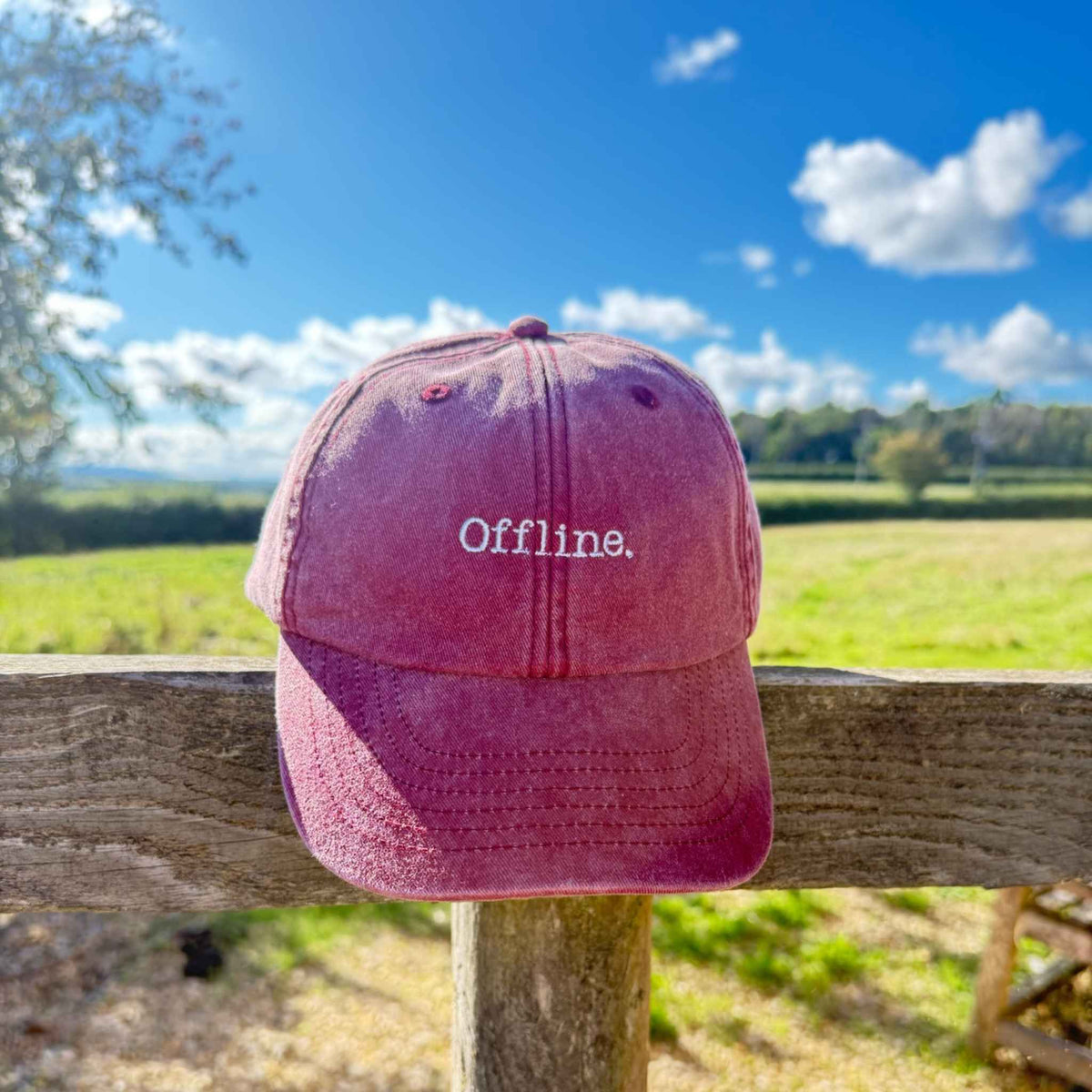 A dusty rose pink baseball cap with 'Offline.' embroidered in white on the front, resting on a wooden fence with a blurred green field and blue sky in the background.