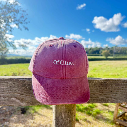 A dusty rose pink baseball cap with 'Offline.' embroidered in white on the front, resting on a wooden fence with a blurred green field and blue sky in the background.