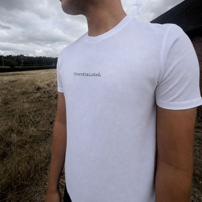 A man wearing a white crew-neck t-shirt with the word 'Overstimulated.' printed in black typewriter font on the chest, photographed outdoors in a field under a cloudy sky.