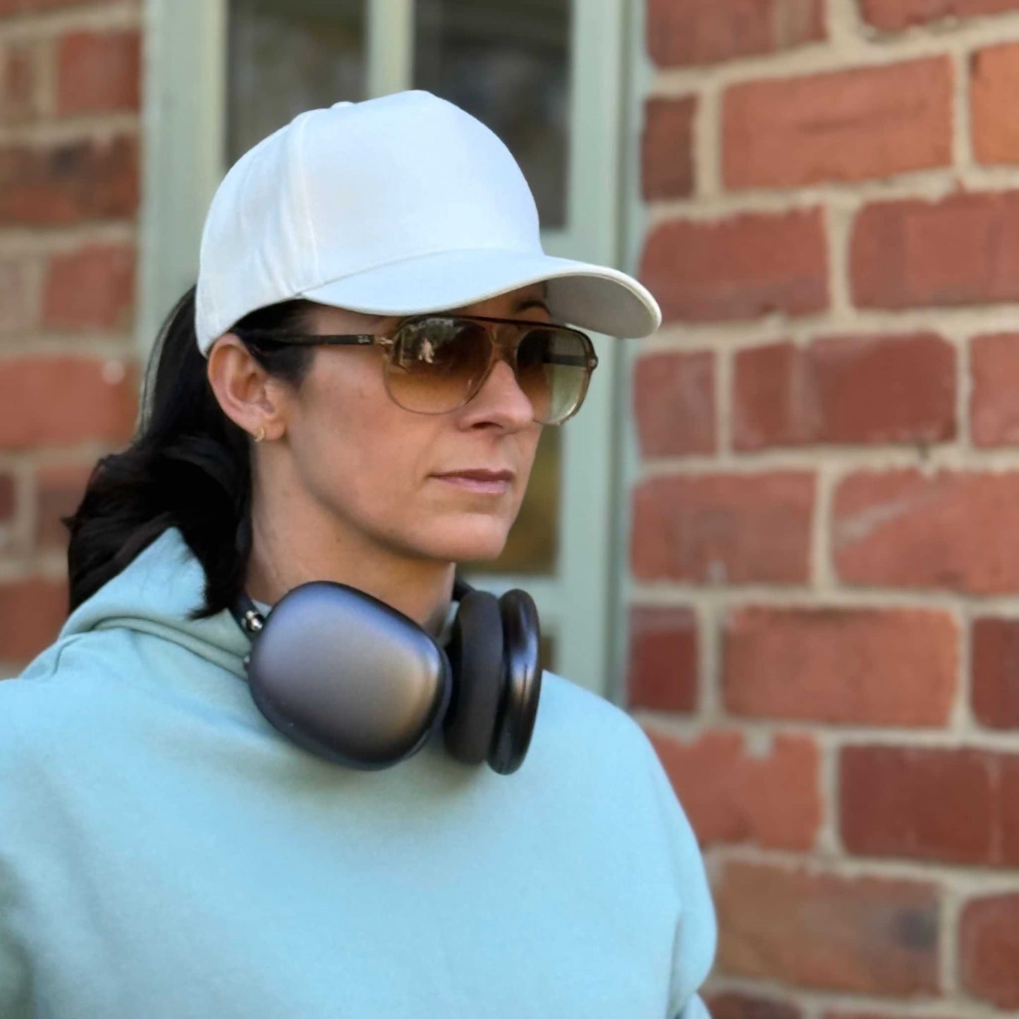 Person wearing a white cap and sunglasses with headphones around their neck, standing in front of a brick wall.