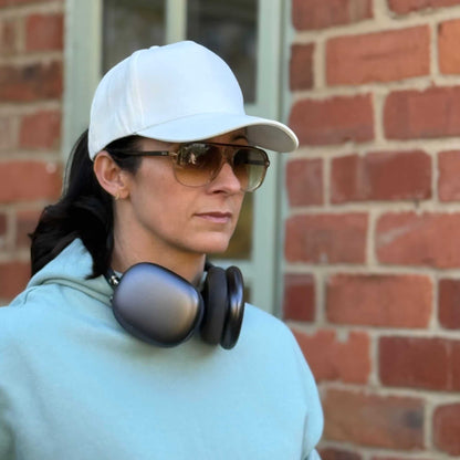 Person wearing a white cap and sunglasses with headphones around their neck, standing in front of a brick wall.