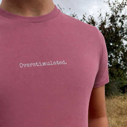 A close-up of a person wearing a dusty rose pink t-shirt with the word 'Overstimulated.' printed in white typewriter font on the chest, set against an outdoor background of trees and dry grass.