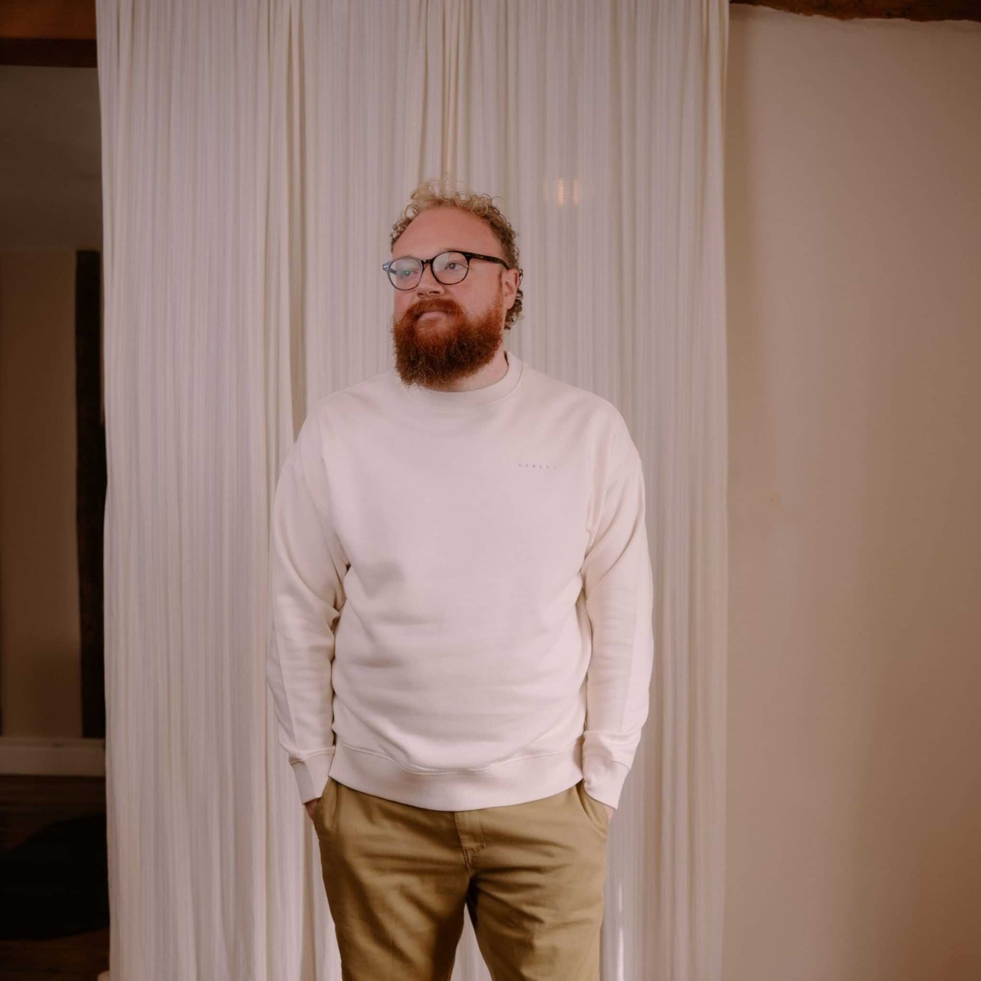 Man wearing a white sweater and beige pants standing in front of a curtain.