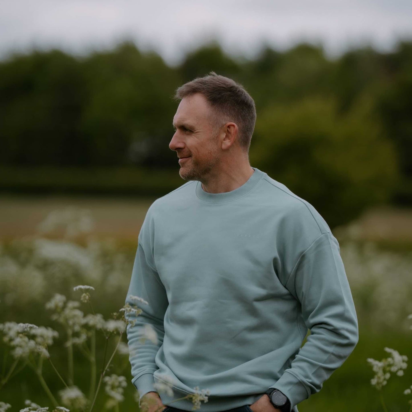 Man in a light blue sweater standing in a field with trees in the background