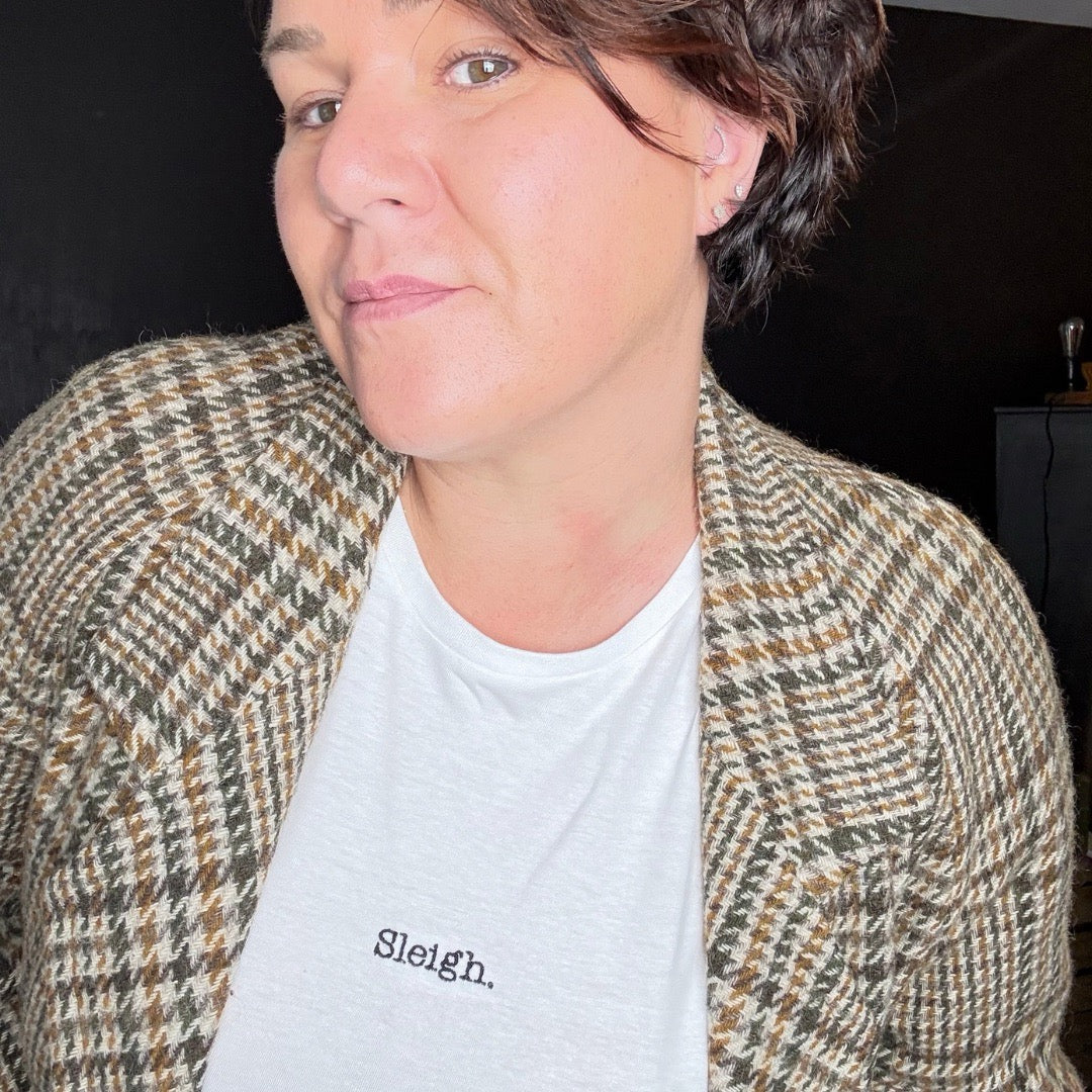 A close-up shot of a person wearing a white crewneck t-shirt with the word 'Sleigh.' printed in black on the chest, layered under a brown and beige houndstooth patterned blazer.