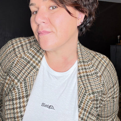 A close-up shot of a person wearing a white crewneck t-shirt with the word 'Sleigh.' printed in black on the chest, layered under a brown and beige houndstooth patterned blazer.