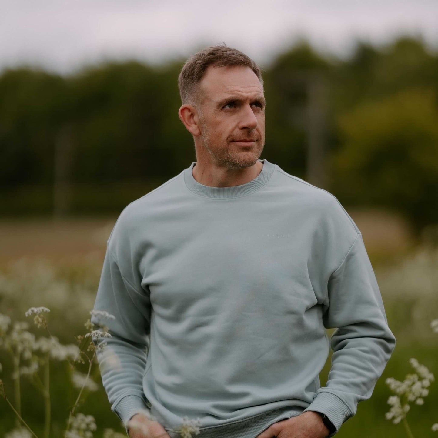 Man wearing a light blue sweater standing in a field with trees in the background