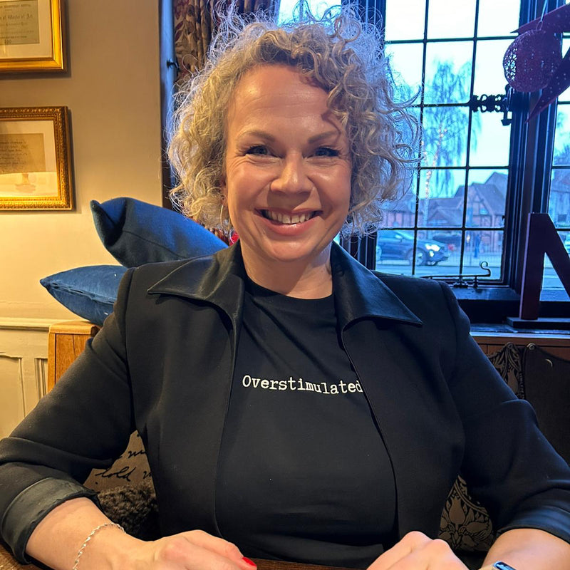 Woman sitting at a table with a window and framed picture in the background wearing overstimulated t-shirt