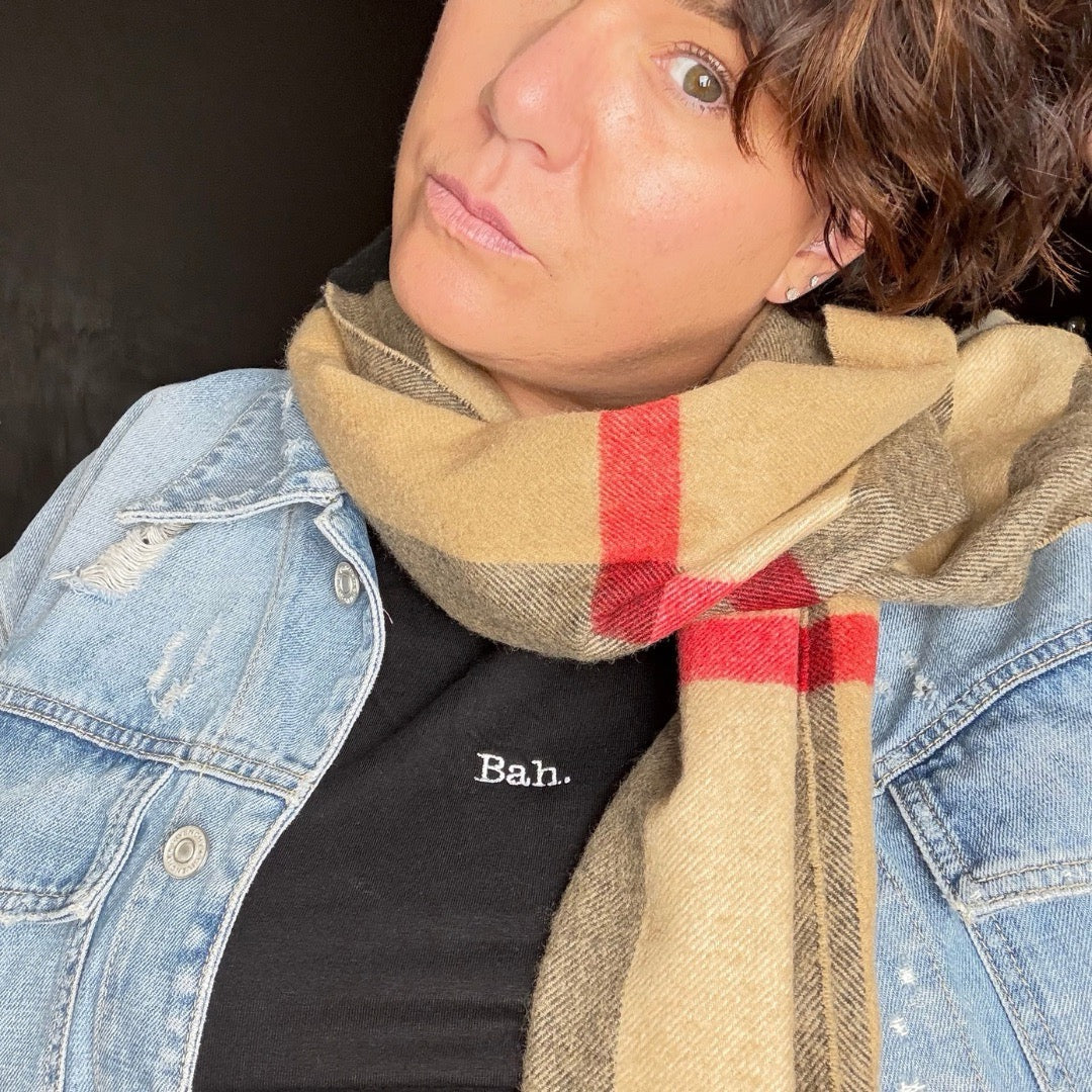 A close-up lifestyle shot of a person wearing a classic beige and red plaid scarf, A black short-sleeve crewneck t-shirt with the word 'Bah.' A christmas t-shirt, women's fitted t-shirt.