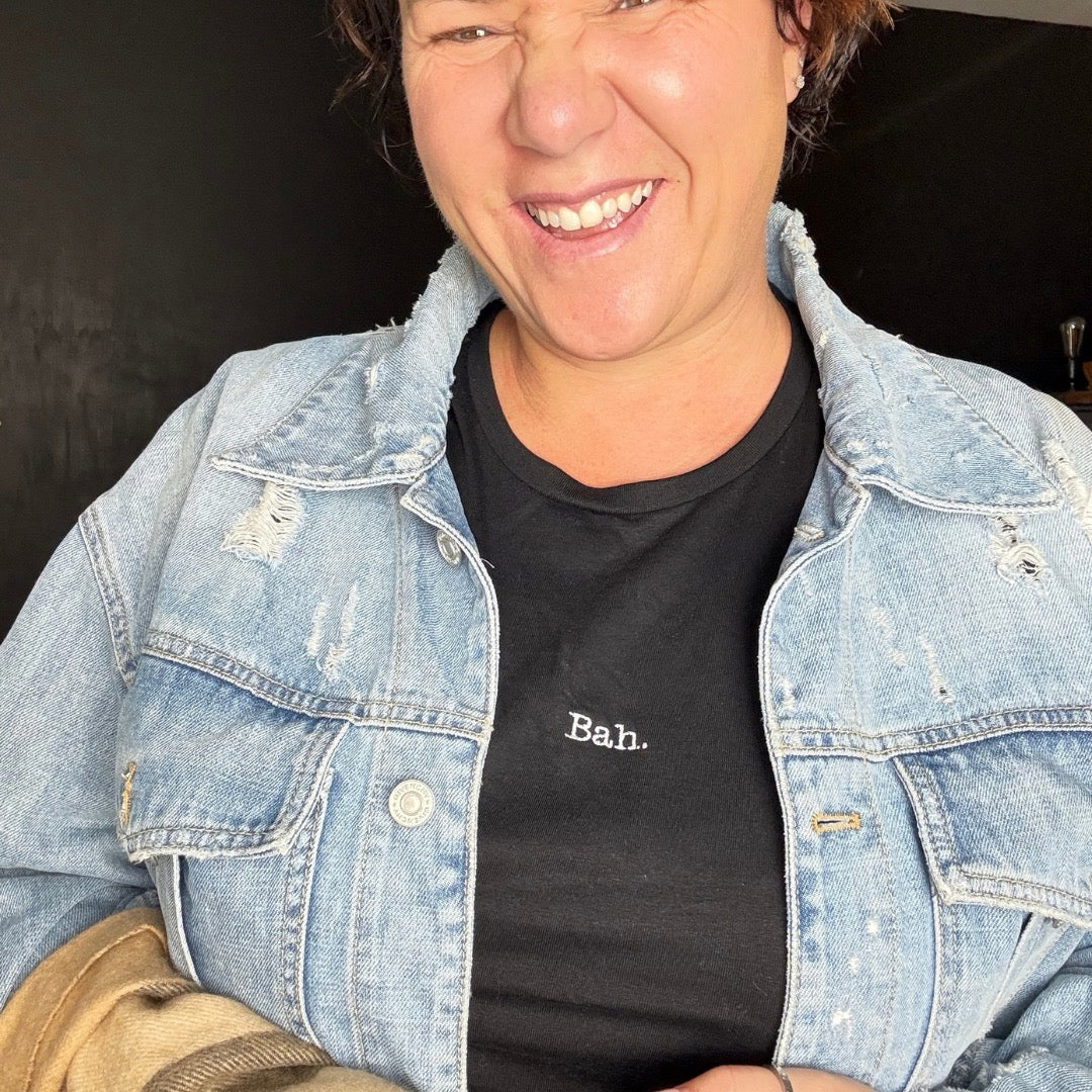 A smiling woman wearing a light-wash distressed denim jacket over a black t-shirt with the word 'Bah.' embroidered in A black short-sleeve crewneck t-shirt with the word 'Bah.' A christmas t-shirt, women's fitted t-shirt.
