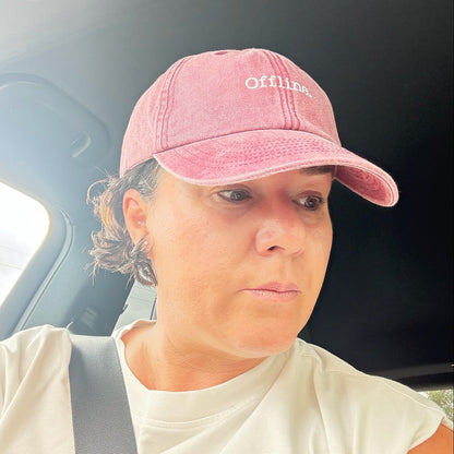 A woman wearing a vintage red baseball cap with offline written onand a white t-shirt sitting inside a car, with the seatbelt across her chest.