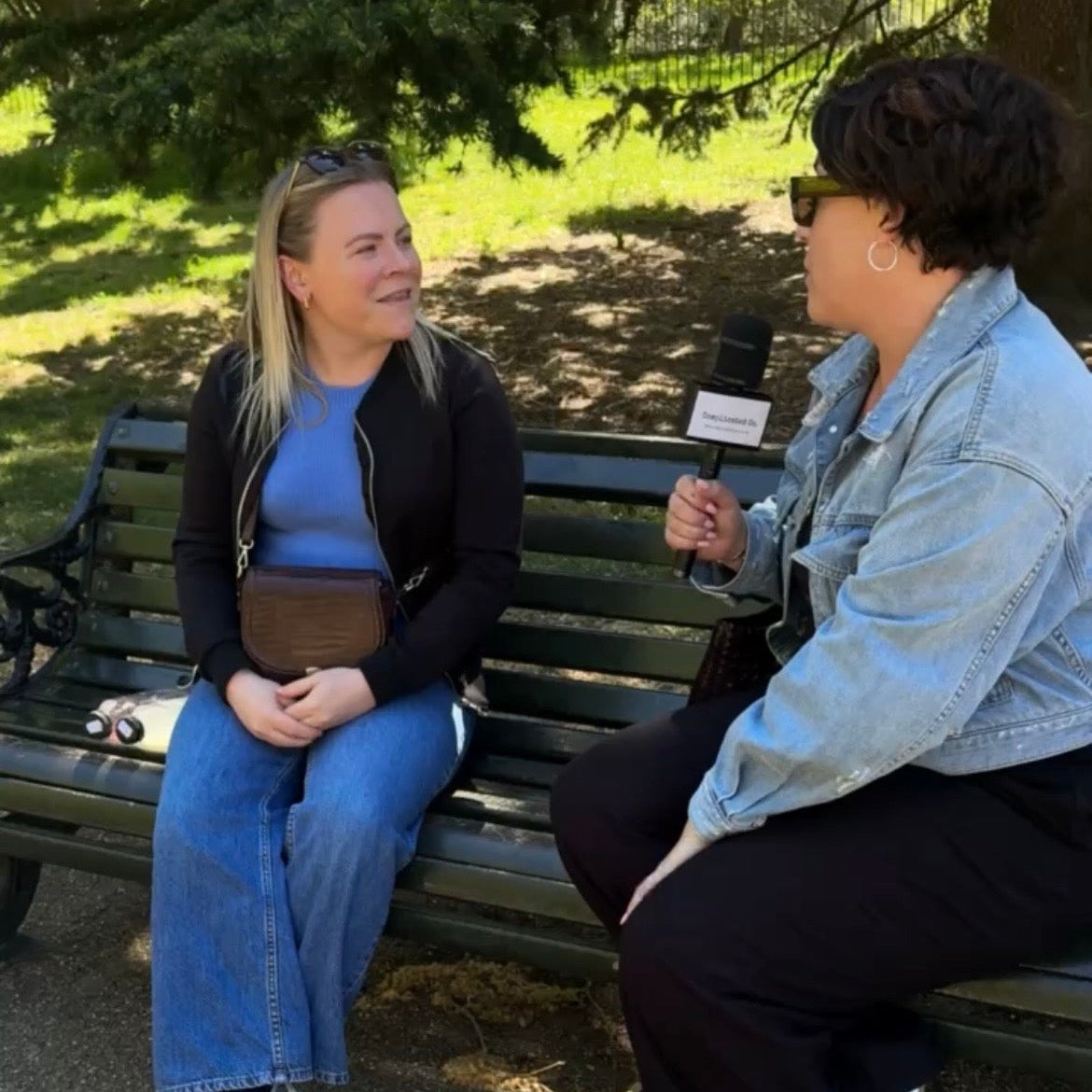 Two women sitting on a park bench, one holding a microphone, in an outdoor setting.