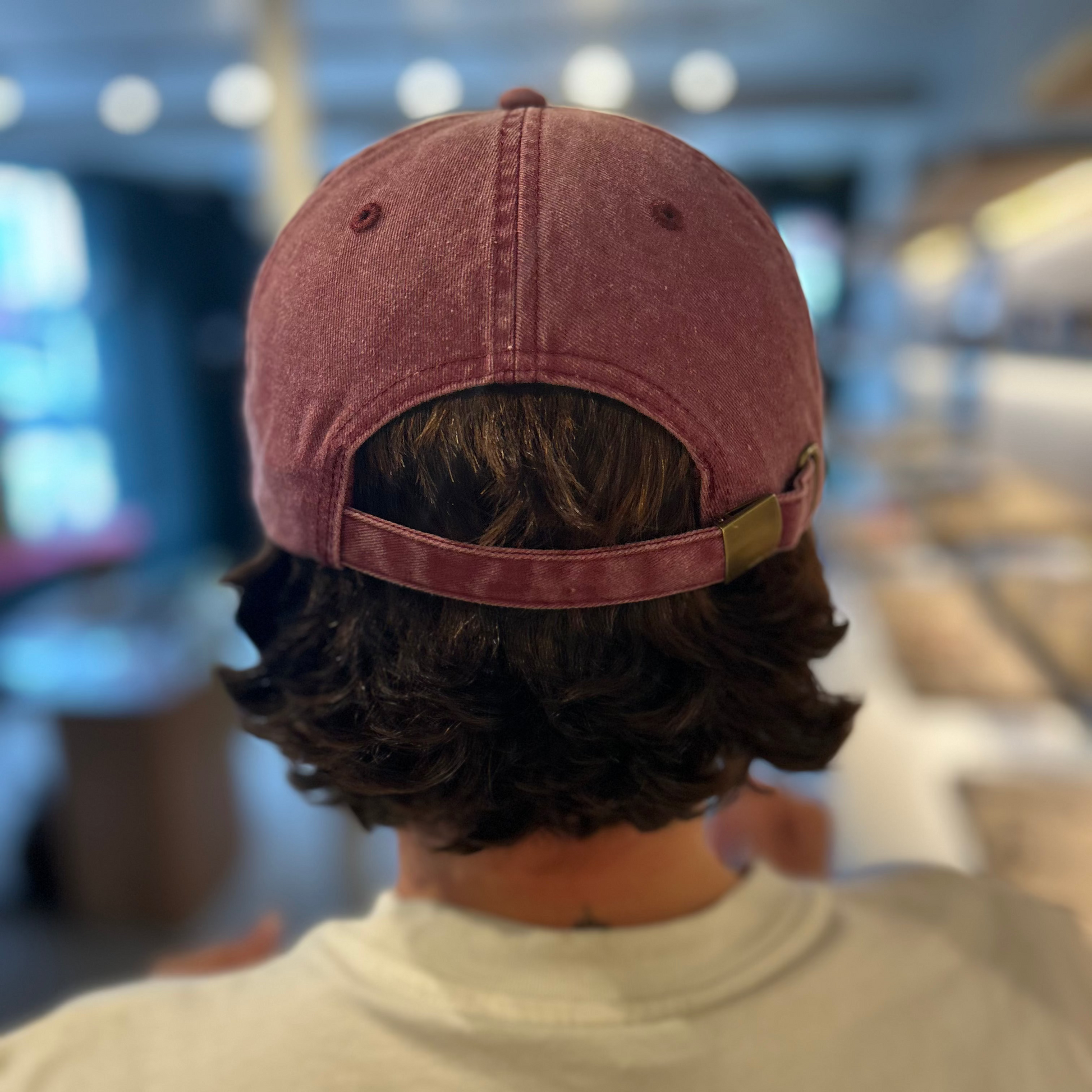 A person viewed from behind wearing a vintage red baseball cap with the strap adjusted, in a blurred indoor retail environment.
