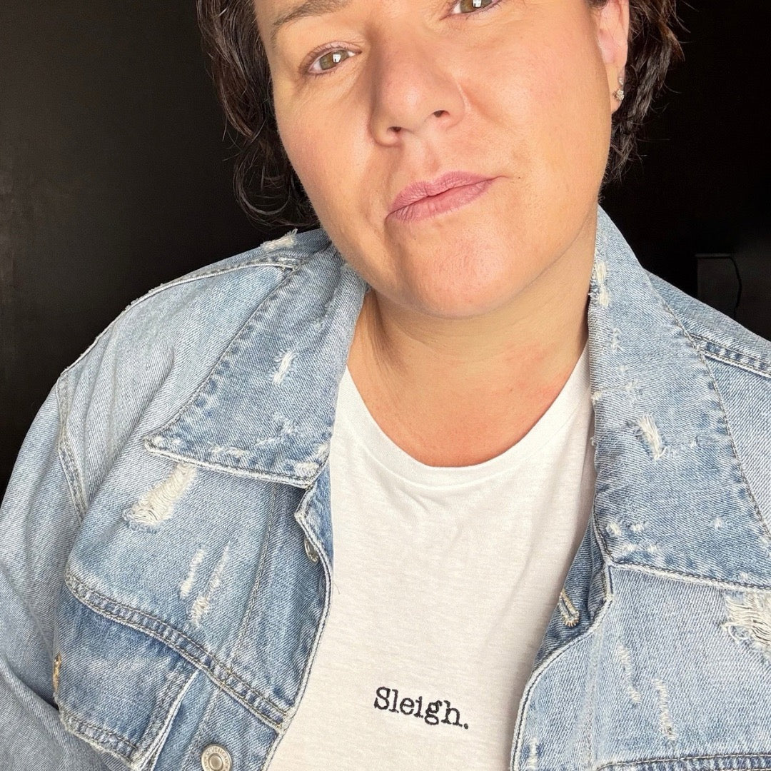 A close-up shot of a person wearing a light-wash, distressed denim jacket over a white crewneck t-shirt with the word 'Sleigh.' printed in black on the chest.