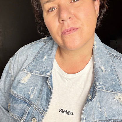 A close-up shot of a person wearing a light-wash, distressed denim jacket over a white crewneck t-shirt with the word 'Sleigh.' printed in black on the chest.