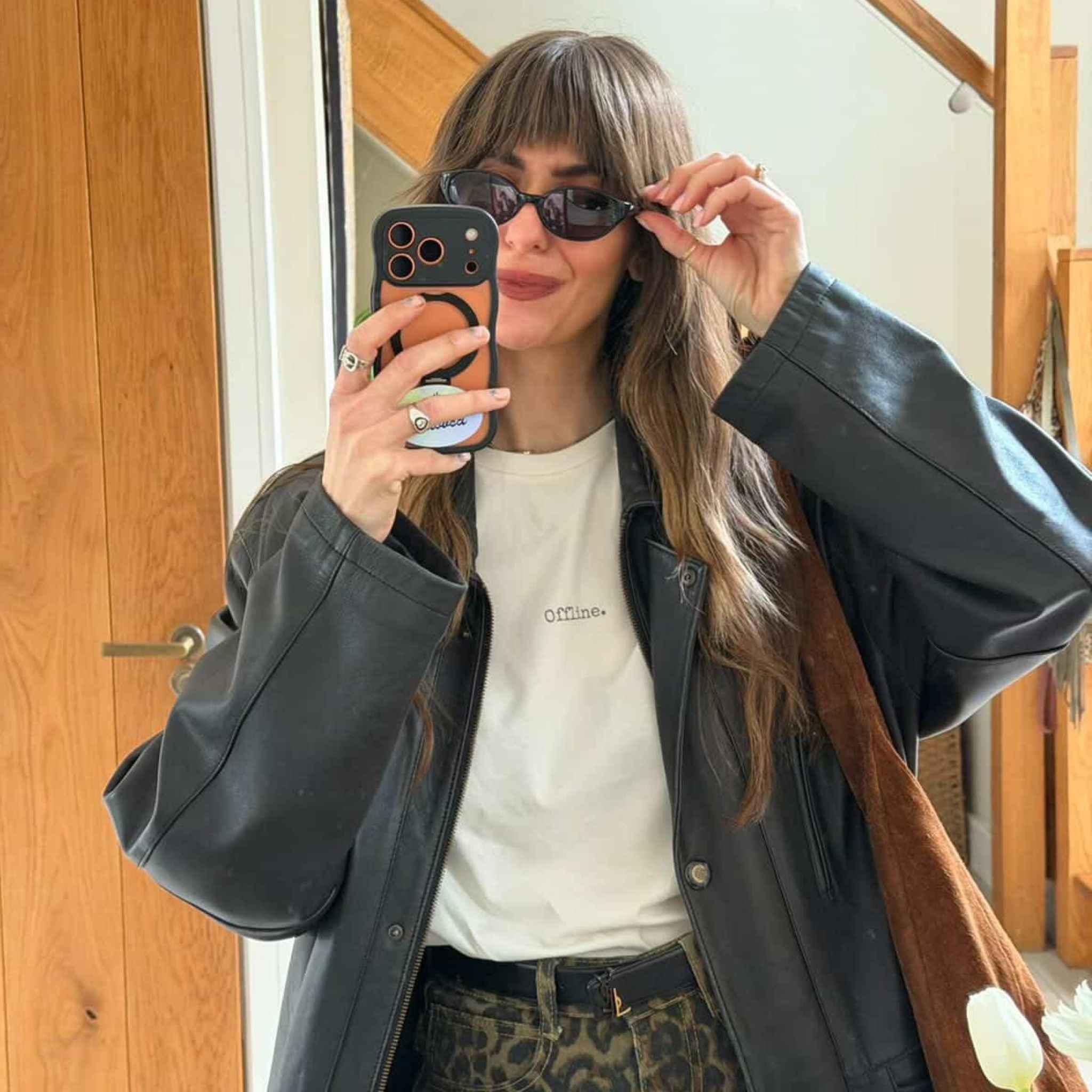 Woman taking a mirror selfie wearing sunglasses, a black jacket, white shirt, and patterned pants.