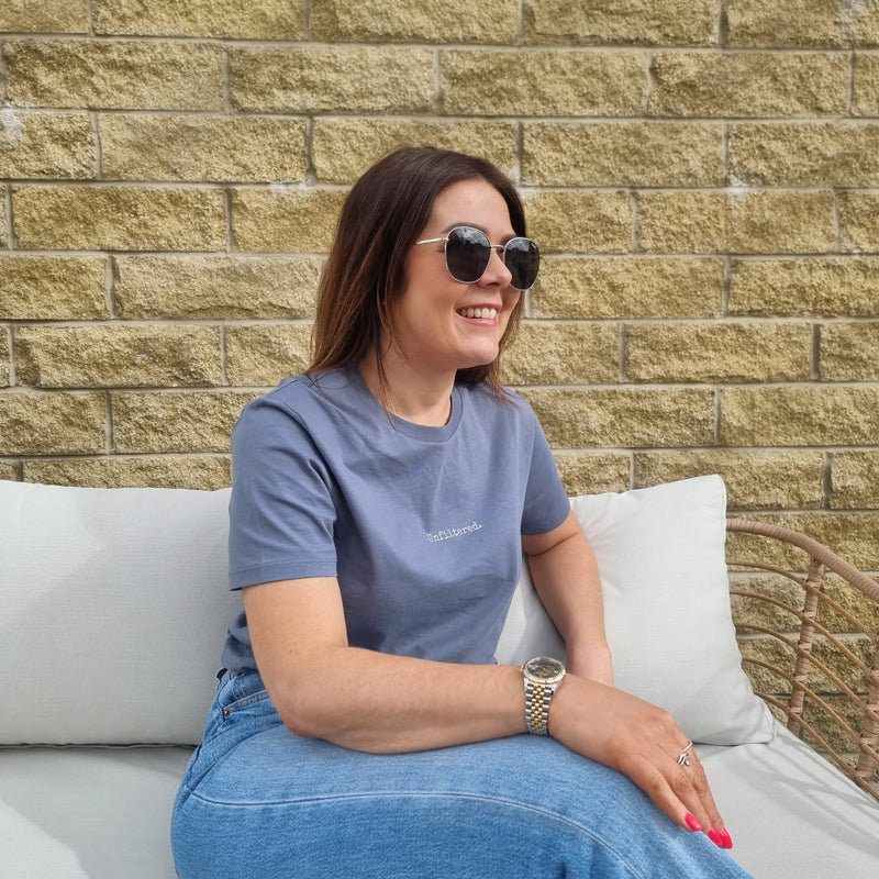 Woman sitting on a white couch wearing sunglasses and a blue t-shirt against a brick wall.