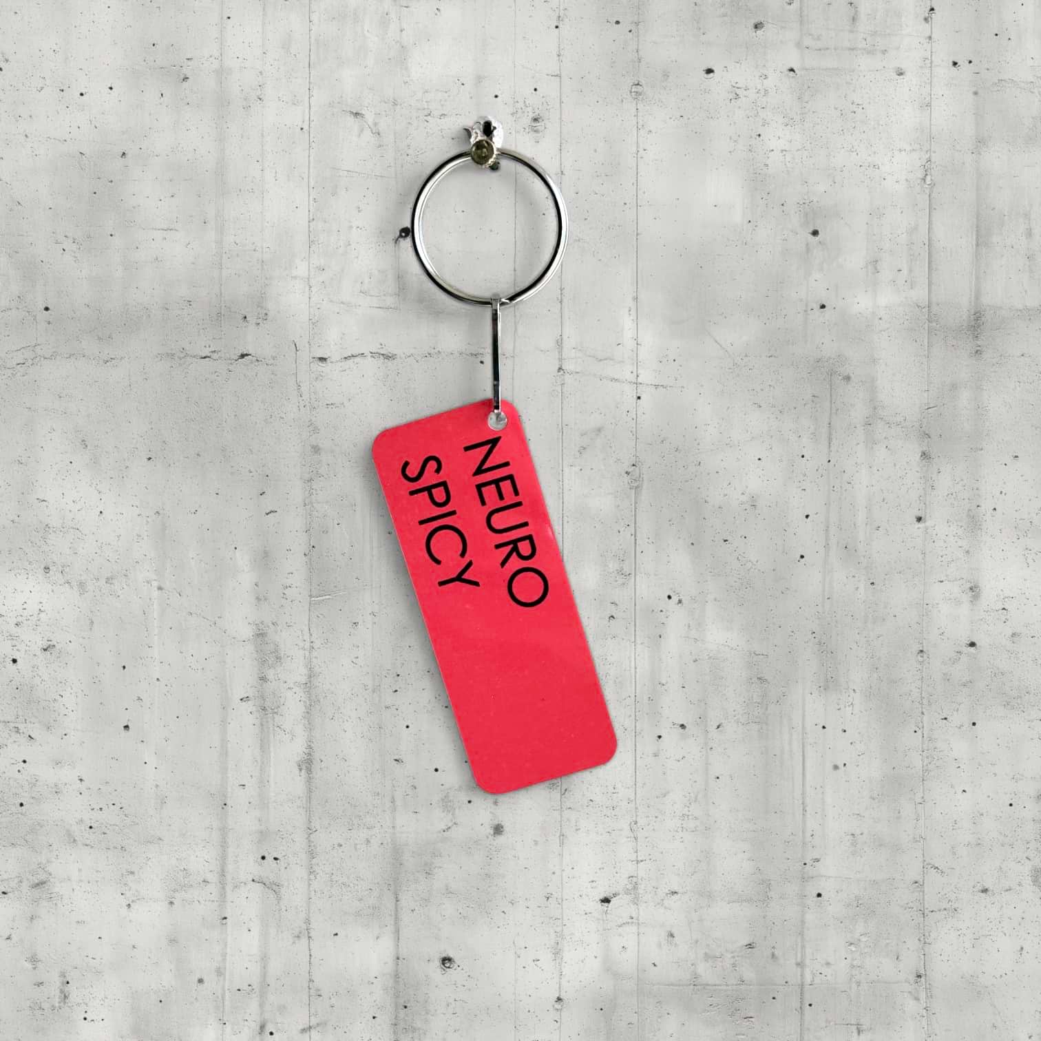 Red keychain tag with 'NEURO SPICY' text on a concrete surface