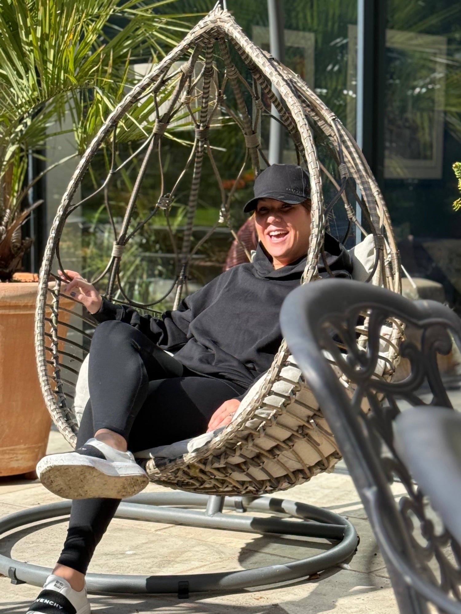 Person sitting in a hanging egg chair in a black hoodie and cap outdoors with plants in the background
